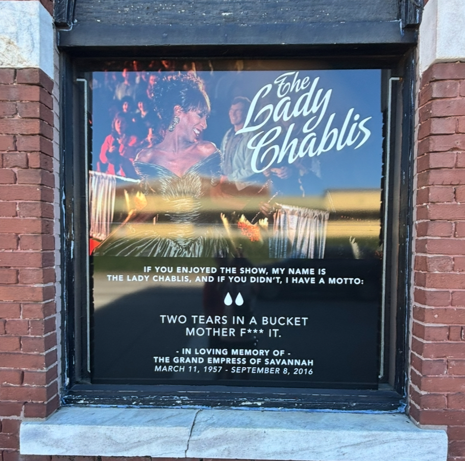 Poster of The Lady Chablis displayed in a window at Club One, visible from the street. She wears a shoulder-baring black gown with curled hair styled in a chignon, smiling with a clutch in her left hand. An audience is visible behind her. The poster includes a tribute: “If you enjoyed the show, my name is The Lady Chablis, and if you didn’t, I have a motto: Two tears in a bucket mother f** it. In loving memory of The Grand Empress of Savannah. March 11, 1957–September 8, 2016.