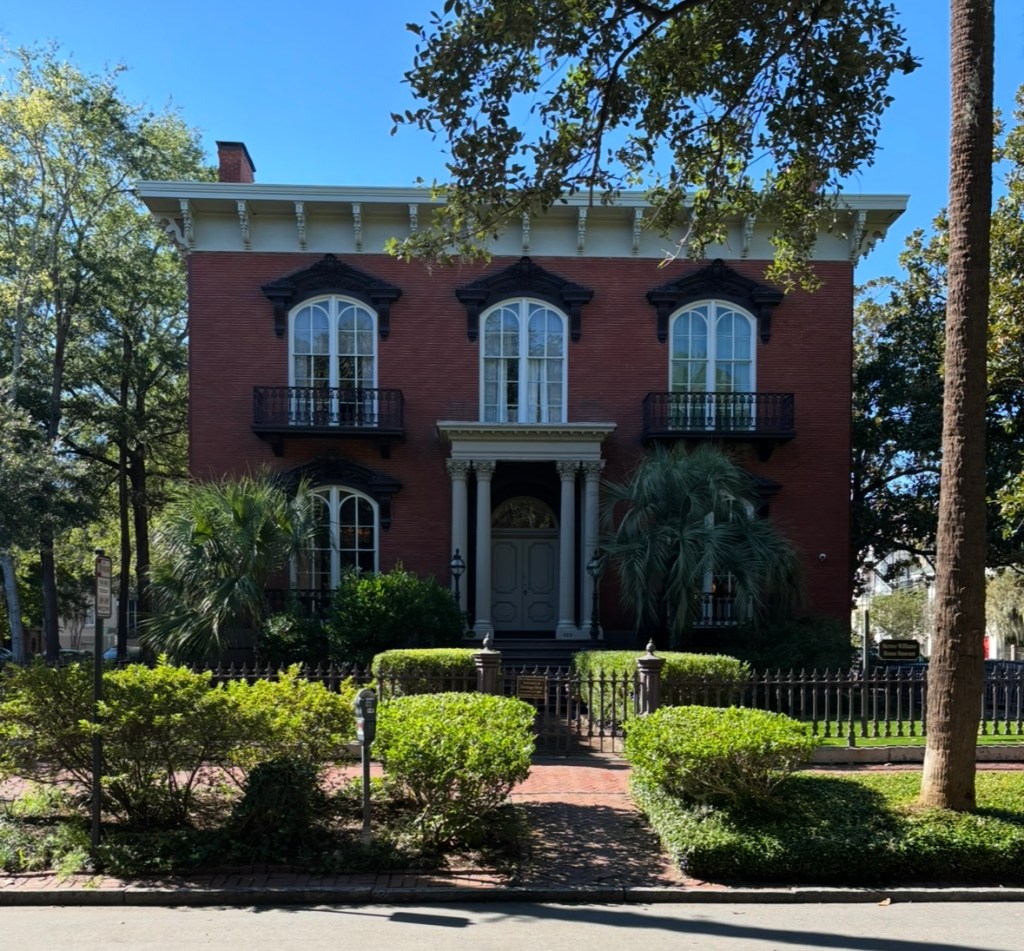 Mercer House Museum, a red brick Italianate mansion with cream-colored columns and a black wrought iron fence. Surrounded by lush greenery and manicured landscaping, the historic home stands as a striking example of 19th-century Savannah architecture.